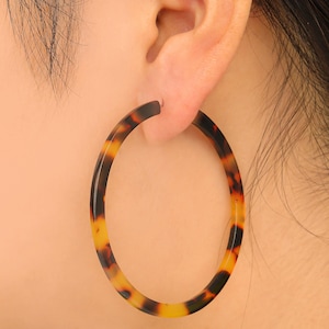 May include: A large, oval-shaped hoop earring with a tortoiseshell pattern. The earring is worn on the ear, showcasing its warm tones of brown, black, and yellow. The earring's design is a classic and versatile accessory.