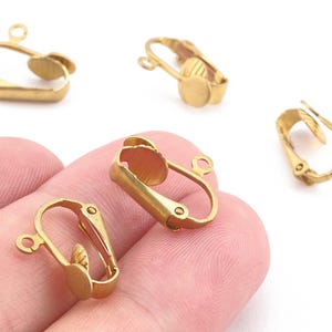 Könnte beinhalten: Mehrere goldfarbene Ohrring-Clip-Verschlüsse werden gezeigt. Jeder Verschluss hat eine Schlaufe zum Anbringen eines Anhängers und einen Klappverschluss mit rundem Polster. Ideal für die Herstellung individueller Ohrringe.