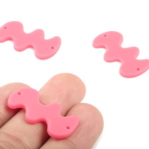 Könnte beinhalten: Drei rosa, wellenförmige Perlen mit jeweils zwei Löchern. Die Perlen haben eine einfarbige, hellrosa Farbe und eine glatte Textur. Eine Perle wird in der Hand einer Person gehalten. Diese Perlen eignen sich für die Schmuckherstellung und das Basteln.