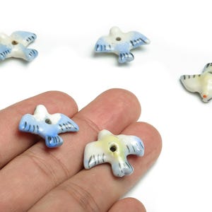 Peut inclure: Petites perles en céramique en forme d'oiseaux, dans des tons de bleu, blanc et jaune. Les perles ont un trou pour l'enfilage et sont disposées sur un fond blanc. Les oiseaux ont des motifs détaillés d'ailes et de queue.