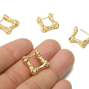 以下が含まれることがあります： 金色の正方形フープピアス。テクスチャ加工された有機的なデザイン。ピアスはポスト留め式で、白い背景に表示されています。ジュエリーはおそらく金属製です。