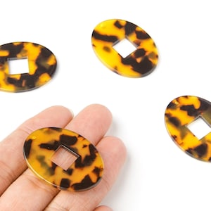 Peut inclure: Perles ovales à motif écaille de tortue avec une découpe carrée au centre. Les perles sont un mélange de jaune et de marron foncé, et l'une d'elles est tenue dans une main. Ces perles sont idéales pour la création de bijoux.