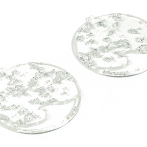 Może przedstawiać: Dwa okrągłe, przezroczyste ozdoby akrylowe z wzorem srebrnych błyskotek. Ozdoby mają otwór na górze do zawieszenia.