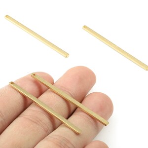 May include: Gold-coloured metal bar pendants with a small hole at one end. The pendants are rectangular and thin, with a matte finish. Several bars are visible, some held in a hand, others scattered on a white background. These are jewellery making supplies.