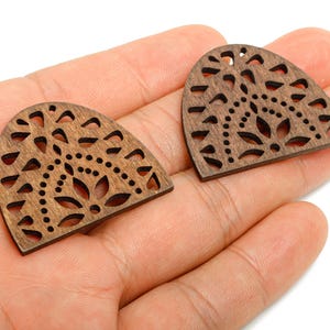 May include: Two wooden, semi-circular pendants with intricate cut-out designs. The pendants are a warm brown color and feature floral and geometric patterns. Each has a small hole at the top for stringing. These are ideal for jewelry making.
