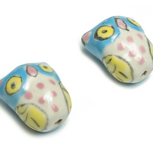 Könnte beinhalten: Zwei Keramik-Eulenperlen mit blauen, gelben und rosa bemalten Details. Die Perlen sind weiß mit einem Loch in der Mitte.