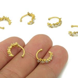 Può includere: Polisini per orecchie color oro con gemme chiare a taglio quadrato. I polsini per orecchie sono disposti a forma di semicerchio, con un design curvo che si avvolge attorno all'orecchio. I gioielli sono esposti su sfondo bianco.