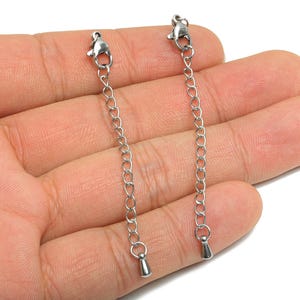 May include: Two silver-colored chain extenders with lobster claw clasps and small cone-shaped end pieces. The extenders are held in a hand, showcasing the jewelry findings. These are likely used for necklace or bracelet adjustments.