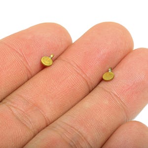 Puede incluir: Dos pequeños colgantes redondos de color dorado con un lazo en la parte superior se muestran en los dedos de una persona. Los colgantes son pequeños, con un diámetro de aproximadamente 3 mm. El fondo es blanco.