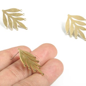 Peut inclure: Composants de boucles d'oreilles en forme de feuille de couleur or. Chaque composant présente un motif de feuille stylisé avec de multiples feuilles ovales et de petites boucles pour la fixation à d'autres éléments de bijoux. Une boucle d'oreille est tenue dans une main.