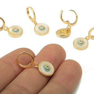 May include: Gold-tone earrings with a white circular pendant and a turquoise center. The earrings have a lever back closure. The design features a small turquoise and crystal accent in the center of the pendant.