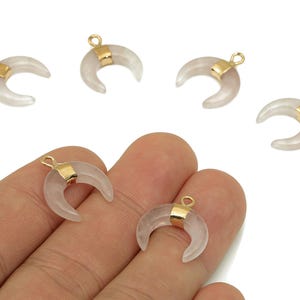 May include: Six crescent moon charms. The charms are a translucent pink with gold-colored metal accents. Two charms are held in a hand. The charms are likely for jewelry making or crafting.