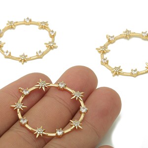 May include: Gold-colored metal charm with a circular design. The charm features a series of star-shaped embellishments and clear, round gemstones. The charm is held in a hand against a white background. Jewelry making supplies.