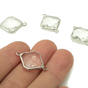 May include: Four clear, faceted, square-shaped crystal beads with silver-colored metal frames and loops. One bead is held in a hand. The beads are on a white background. These beads are suitable for jewelry making.