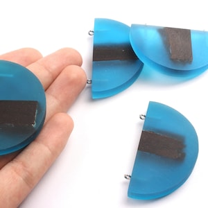 May include: Several blue resin half-circle pendants with a dark rectangular inlay. Each pendant has a small silver-colored loop at the top for attaching to a chain or other jewelry component. The pendants are displayed on a white background.