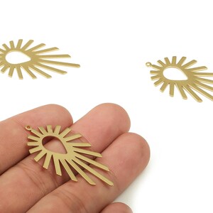 Puede incluir: Encantos de metal dorado con forma de sol con un centro en forma de lágrima y líneas radiantes. Un encanto se sostiene en una mano, mientras que otros dos se muestran sobre un fondo blanco. Estos encantos son adecuados para la fabricación de joyas.