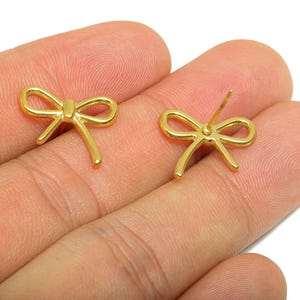 Pode incluir: Um par de brincos de botão dourados em forma de laço. Um brinco é mostrado com o pino e o outro sem ele. Os brincos são pequenos e delicados, com um acabamento polido. Os brincos são exibidos em um tom de pele claro.