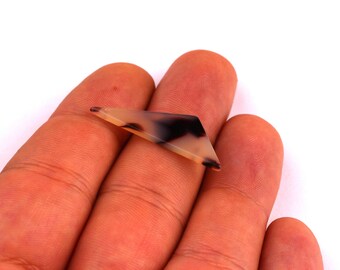 Puede incluir: Un pequeño elemento decorativo de forma geométrica con un patrón de carey en tonos beige, negro y crema. El objeto se sostiene en una mano sobre un fondo blanco. La forma es angular con una superficie plana.