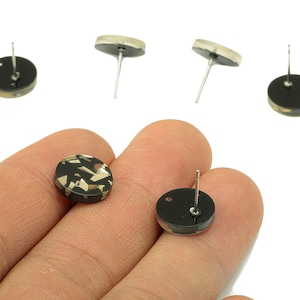 Pode incluir: Brincos de botão redondos pretos e dourados com um design geométrico. Os brincos têm formato circular com fecho de pino. Vista de perto dos brincos.