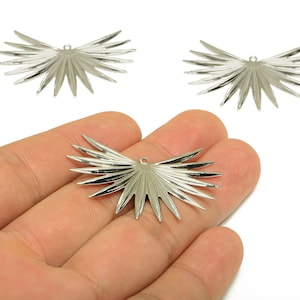 May include: Three silver-colored metal charms in a fan or wing shape. One charm is held in a hand, while two others are positioned above. Each charm has a small hole at the top for stringing.
