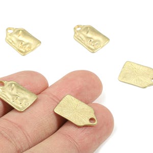 Op de afbeelding: Verschillende kleine, goudkleurige metalen tag bedels met een getextureerd oppervlak en een gat om te rijgen. Eén bedel wordt in een hand gehouden, terwijl de andere verspreid zijn op een witte achtergrond. Deze bedels zijn geschikt voor het maken van sieraden.