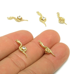 May include: Gold-coloured snake charms with a clear gemstone accent. The charms are in a coiled snake design, with a loop at the top for attaching to jewellery. Several charms are scattered on a white background, with two held in a hand.