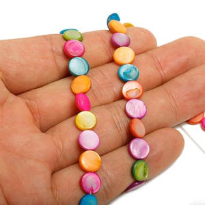 May include: A strand of colorful, iridescent shell beads in shades of pink, orange, yellow, blue, and green. The beads are round and flat, and are held in a hand against a white background. The beads are approximately 0.5 inches in diameter.