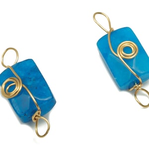 Puede incluir: Dos colgantes de piedra azul con alambre dorado envuelto alrededor. El alambre está retorcido en forma de espiral.