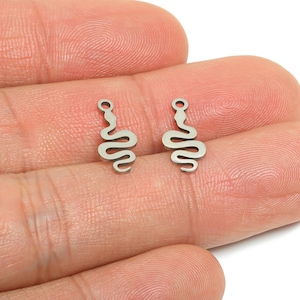 Puede incluir: Dos pequeños colgantes plateados con forma de serpiente. Cada colgante tiene un lazo en la parte superior y un diseño de serpiente estilizado. Los colgantes se muestran sobre un fondo color piel.