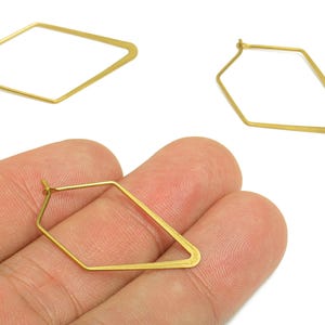 Op de afbeelding: Drie goudkleurige, geometrische oorbellen. Twee oorbellen worden apart weergegeven, terwijl de derde in een hand wordt gehouden. De oorbellen hebben een unieke, hoekige vorm, met een haaksluiting.