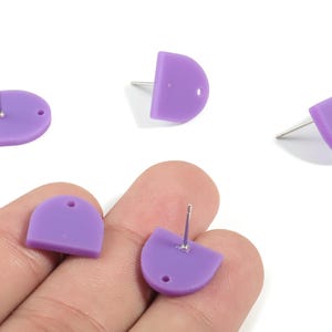 May include: A selection of lavender-coloured, semi-circular stud earrings. Each earring has a post for piercing and a small hole. The earrings are displayed on a white surface, with some held in a hand.