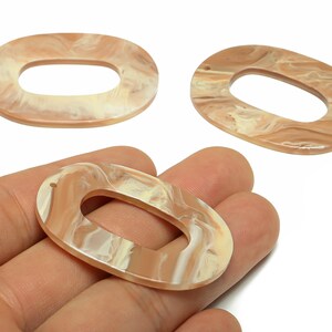 Op de afbeelding: Drie ovale harskralen met een gemarmerd patroon in beige en crème. Eén kraal wordt in een hand gehouden. Deze kralen zijn geschikt voor het maken van sieraden en knutselprojecten.