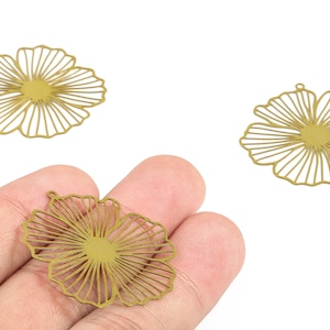 Peut inclure: Trois breloques en forme de fleur ajourée de couleur dorée. L'une est tenue dans une main, tandis que deux autres sont positionnées à proximité. Les breloques ont un design radial détaillé, avec une petite boucle en haut pour suspendre. Idéal pour la création de bijoux.