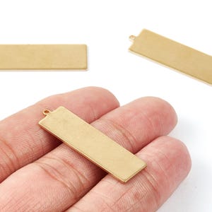 May include: Three rectangular, gold-colored metal pendant blanks. One is held in a hand, while two others are displayed separately. Each blank has a small loop at one end for attaching to a chain or other jewelry component.