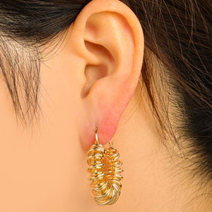 May include: Gold-colored hoop earrings with a unique coiled design. The earrings are made of a shiny, metallic material and are worn on the ear. The design features multiple loops, creating a textured and eye-catching look. These earrings are a stylish accessory.