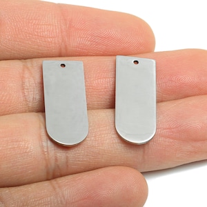 Puede incluir: Dos colgantes rectangulares plateados con fondos redondeados y un pequeño agujero en la parte superior para la fabricación de joyas. Los colgantes se sostienen en una mano sobre un fondo blanco.