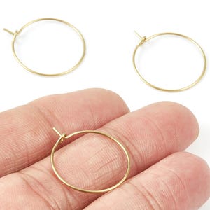 May include: Three gold-colored hoop earrings. One earring is held in a hand, while the other two are on a white surface. The earrings are simple, round, and made of thin metal wire. They are a classic jewelry design.