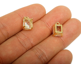 Puede incluir: Dos colgantes rectangulares dorados con piedras claras y facetadas. Un colgante es un rectángulo sólido con un lazo en la parte superior, mientras que el otro tiene un diseño de marco abierto. Los colgantes se muestran en una mano.
