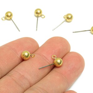 May include: Gold-coloured ball stud earrings with a post and loop. The earrings are small and round, with a shiny finish. Several earrings are scattered on a white background.