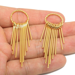 May include: A pair of gold-colored earrings. Each earring features a circular top with a twisted rope design, from which multiple straight, thin bars extend downwards. The earrings are held in a hand.