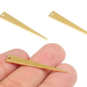 Könnte beinhalten: Drei goldfarbene, längliche Dreiecksanhänger mit einem kleinen Loch oben. Ein Anhänger wird in einer Hand gehalten. Die Anhänger haben eine einfache, geometrische Form, die sich für die Schmuckherstellung oder andere Bastelarbeiten eignet.