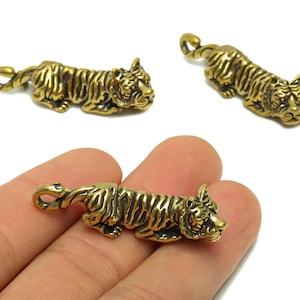 May include: Three small, detailed brass tiger figurines. The tigers are lying down with their tails curled. The brass has a golden hue, and the tigers have striped patterns. One figurine is held in a hand.