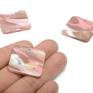 May include: Three square resin pendants with rounded corners. The pendants feature a marbled design in shades of pink, white, and gold. Each pendant has a small hole for stringing.