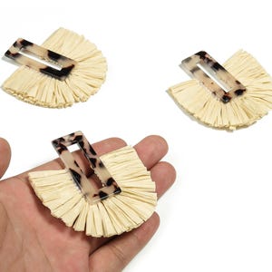 Peut inclure: Trois pinces à cheveux avec un design en éventail de raphia naturel et une pince rectangulaire à motif écaille de tortue. Le raphia est de couleur beige clair, et les pinces sont sur fond blanc.