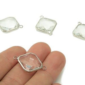 Pode incluir: Berloques conectores de vidro transparente e facetado com molduras de metal prateado. Os berloques têm uma forma geométrica com dois laços para fixar à joalharia. Vários berloques são visíveis, um deles sendo segurado na mão.