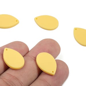 Pode incluir: Pingentes em forma de gota amarelo pálido com um pequeno orifício na parte superior para enfiar. Vários pingentes estão espalhados sobre uma superfície branca, com dois sendo segurados em uma mão. Estes pingentes são adequados para fazer joias.
