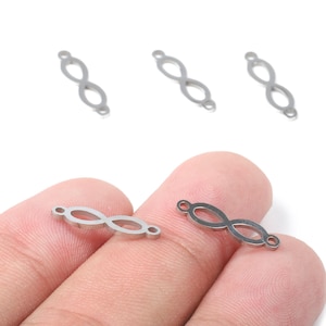 May include: Silver-coloured infinity charm connectors. These small, open-link charms feature a figure-eight design with a loop at each end. The charms are shown on a white background and held by a person's fingers.