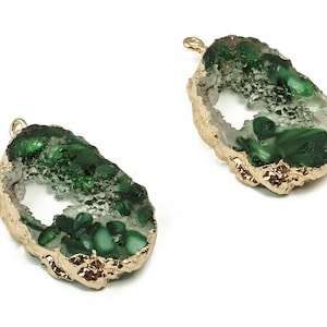 May include: Two gemstone pendants with a gold-colored trim. The pendants have a teardrop shape with a hollow center filled with green and white crystals. The outer edge is a gold color, and each pendant has a small loop at the top.