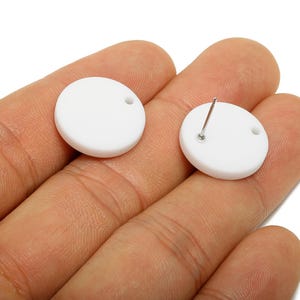 Pode incluir: Dois brincos redondos brancos com um pequeno orifício e um pino de prata. Um brinco é mostrado com o pino preso e o outro sem o pino. Os brincos são segurados na mão de uma pessoa.