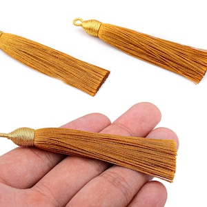 May include: Three golden-brown tassels with a loop at the top for hanging. One tassel is held in a hand. The tassels are made of many thin strands of thread, and the top is wrapped in a gold-colored material.